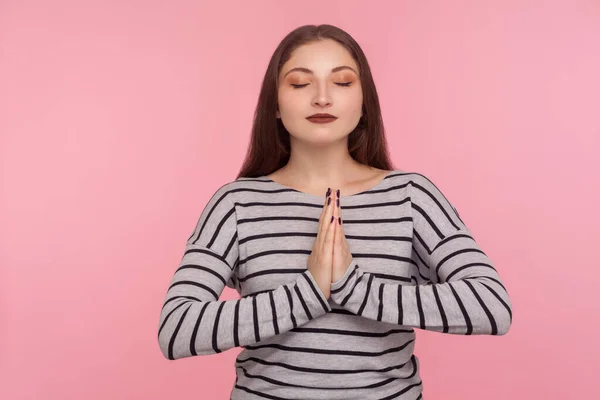 Yoga meditation, inner peace. Portrait of tranquil woman in striped sweatshirt holding hands in namaste gesture and practicing mental health, breath techniques. studio shot isolated on pink background