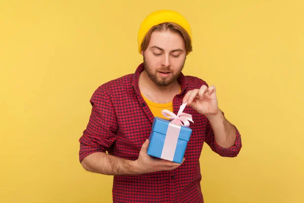 Positive hipster guy in beanie hat and checkered shirt untying bow on gift box, unwrapping present with interest, in anticipation of nice birthday surprise. studio shot isolated on yellow background