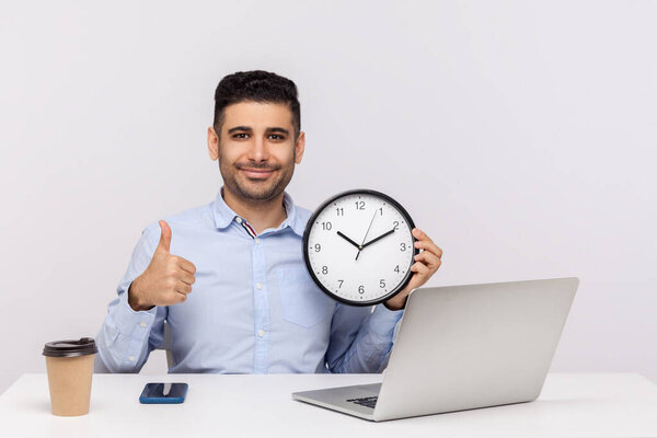 Time management. Cheerful young man employee sitting in office workplace with laptop, holding big clock and showing thumbs up, like gesture, smiling at camera. studio shot isolated on white background