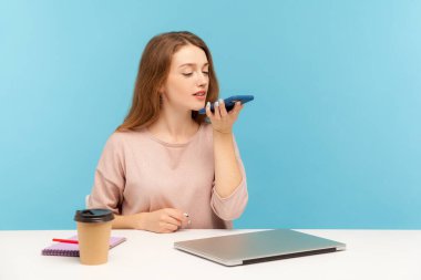 Dijital hoparlör uygulaması. Genç bayan çalışan işyerinde oturuyor ve cep telefonuyla konuşuyor, iş etkinliğini hatırlatmak için ses asistanı kullanıyor. Mavi arka planda kapalı stüdyo çekimleri