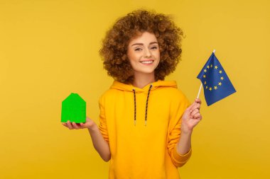 AB 'de gayrimenkul alımı, göç. Şehir tarzı kapüşonlu mutlu kıvırcık saçlı genç kadının portresi. Elinde Avrupa bayrağı ve kağıt ev var. Sarı arka planda izole edilmiş kapalı stüdyo çekimi