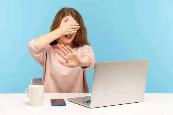 Don't want to look at this. Confused woman employee siting at workplace with laptop and covering eyes with hand, gesturing stop, denial rejection sign. indoor studio shot isolated on blue background