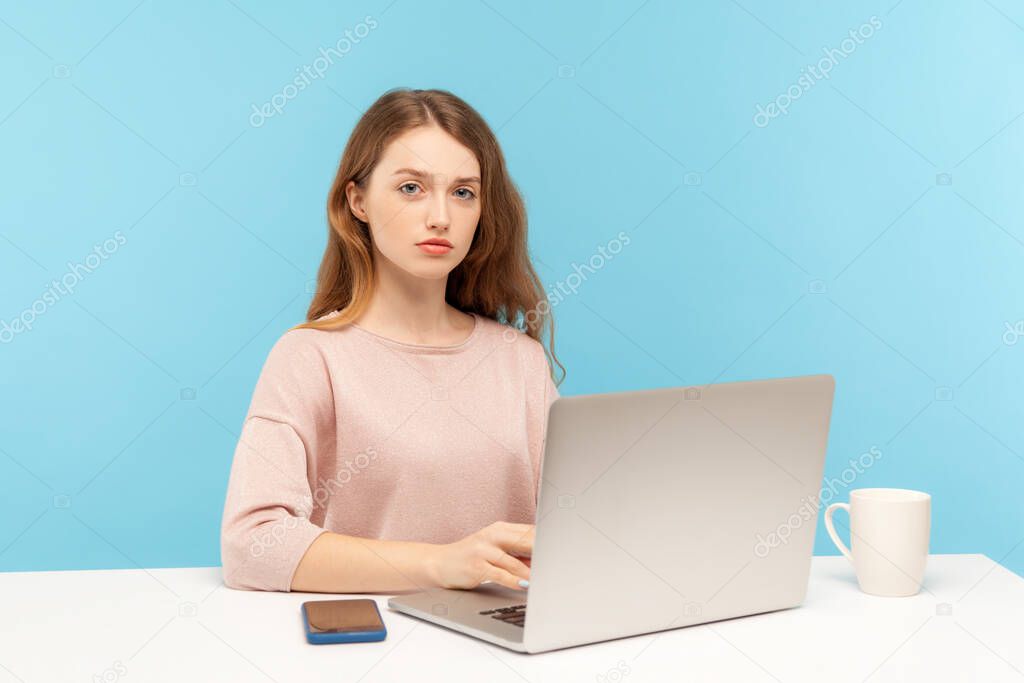 Smart serious young pretty businesswoman in casual clothes looking at camera with responsible confident expression, working on laptop at home office. indoor studio shot isolated on blue background