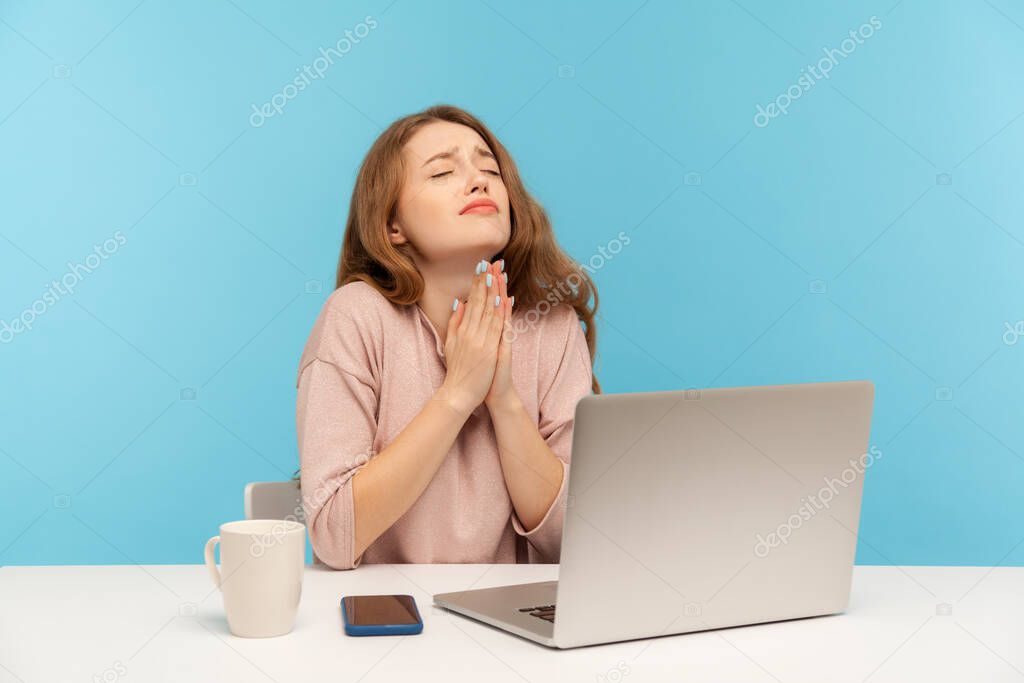 Please, god help! Young woman employee sitting at workplace and keeping eyes closed, praying heartily, pleading with desperate sincere expression. indoor studio shot isolated on blue background