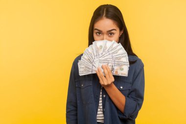 Kot gömlekli mutlu esmer kız portresi dolar banknotlarından, neşeli bir bakışla, para, büyük kar, finansal tasarruflar gösteren. Sarı arka planda izole edilmiş kapalı stüdyo çekimi
