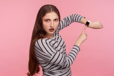 Acele edin! Çizgili eşofman üstü giymiş sinirli bir kadının portresi. Gece geç saatlerde gergin, kol saatini gösteren, endişeli ve son teslim tarihinden endişeli görünüyor. Pembe arka planda kapalı stüdyo çekimleri