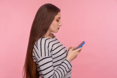 Cep telefonunda yazan, blog okuyan, sosyal ağda sohbet eden, internet iletişimi için mobil uygulama kullanan ciddi özenli genç bir kadının yan görüntüsü. Pembe arka planda kapalı stüdyo çekimleri