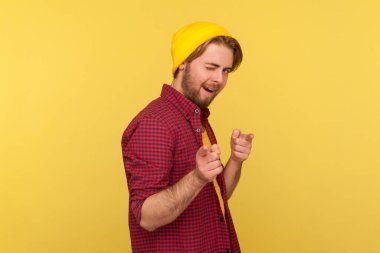 Hey sen! Şapkalı ve damalı gömlekli komik hippi sakallı adam kamerayı işaret ediyor, şakacı bir şekilde göz kırpıyor, kazananı fark ediyor ve mutlu bir şekilde gösteriyor. Stüdyo çekimi sarı arkaplanda izole edildi