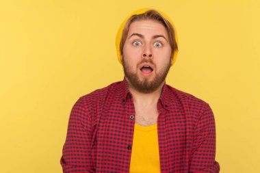 Vay canına, inanılmaz! Şok olmuş bereli ve kareli gömlekli hippi adam kameraya büyük gözlerle bakıyor, ağzı açık, ani haberlere şaşırıyor. Sarı arka planda izole edilmiş kapalı stüdyo çekimi