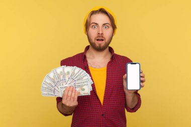Cep telefonu para transferi, online bankacılık. Damalı gömlekli, elinde dolar banknotları ve boş bir cep telefonu olan hippi bir adam reklam için hazırlanıyor, kameraya bakıp şaşkına dönüyor. izole