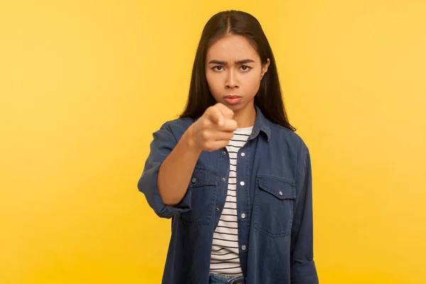 Hey you! Portrait of strict bossy angry girl in denim shirt pointing finger to camera, teaching and scolding, giving serious advice, choosing guilty. indoor studio shot isolated on yellow background
