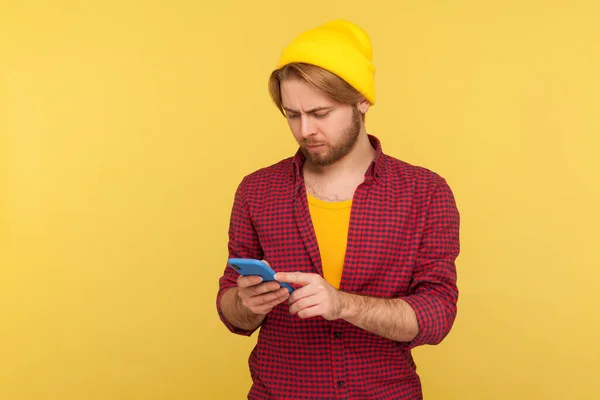 ビーニー帽子のヒップスタートレンディーな男と携帯電話を使用してシャツをチェック テキストメッセージや読書メッセージ ソーシャルネットワークでチャット モバイルアプリケーションを楽しんで 黄色の背景に隔離されたスタジオショット — ストック写真