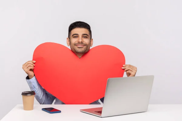 快乐英俊的男人坐在笔记本电脑前 手持大大的红心贺卡 微笑着看着相机 庆祝情人节 在白色背景下隔离的室内拍摄 — 图库照片