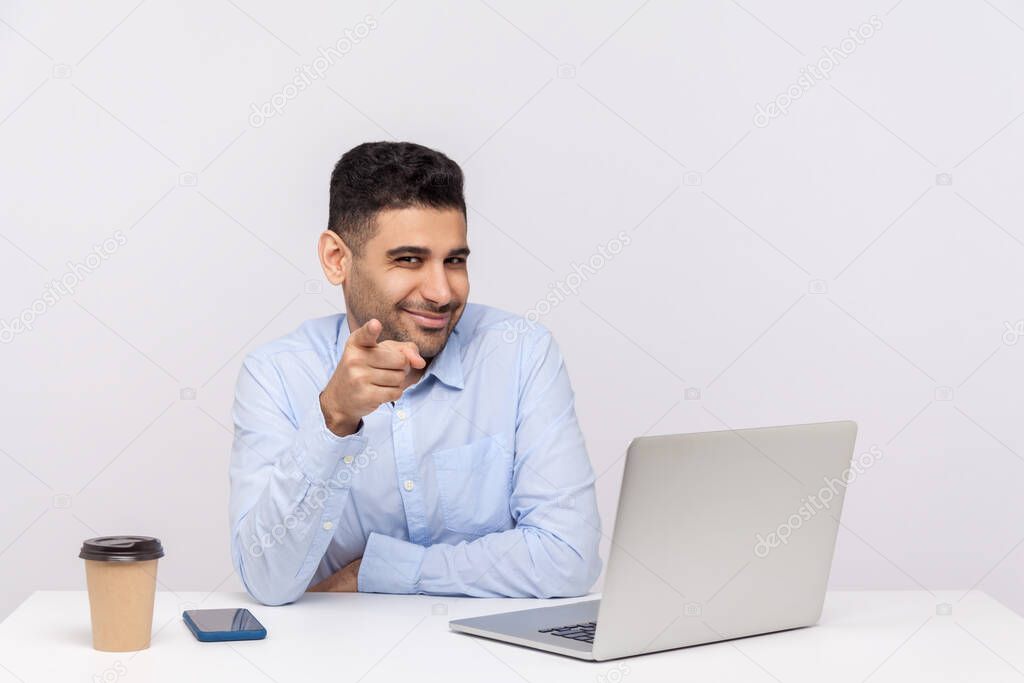 Glad smiling businessman sitting office workplace with laptop on desk, pointing finger to camera, looking pleased happy, choosing lucky coworker. indoor studio shot isolated on white background