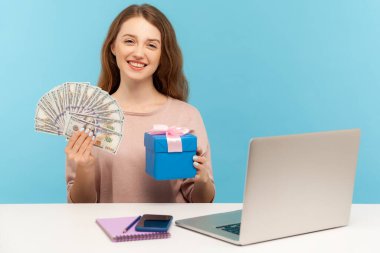 İş yerinde elinde dolar banknotları ve hediye kutusuyla mutlu bir kadın dişlek tebessümle kameraya bakıyor, hediyenin, nakitin ve ikramiyenin tadını çıkarıyor. Mavi arka planda kapalı stüdyo çekimleri