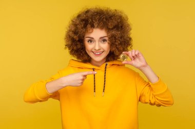 Kripto para, dijital para. Şehir tarzı kapüşonlu mutlu kıvırcık saçlı kadının portresi altın bitcoin 'i gösteriyor ve gülümsüyor, btc sikkeye işaret ediyor. Sarı arka planda izole edilmiş kapalı stüdyo çekimi