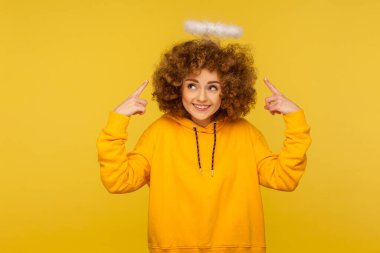 Portrait of charming angel with halo, kind curly-haired hipster woman pointing at saint nimbus over her head and smiling with obedient modest expression. studio shot isolated on yellow background clipart