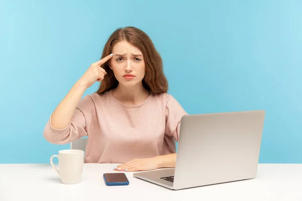 This is crazy silly idea! Displeased woman sitting at workplace with laptop and making stupid gesture, finger near head, teasing senseless idiotic mind. indoor studio shot isolated on blue background
