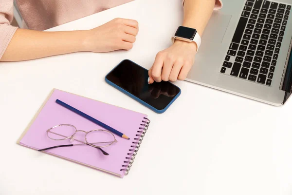 Top view, closeup of woman journalist, businessperson with smart wristwatch sitting at workplace, laptop, phone and paper notebook with glasses on desk. indoor studio shot isolated on blue background