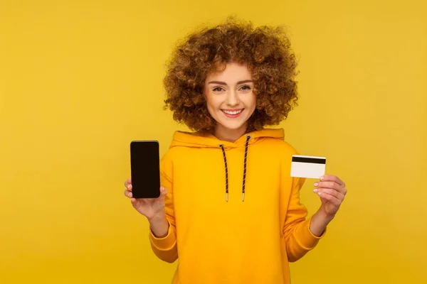 Online banking app, electronic money. Portrait of happy curly-haired woman in urban style hoodie showing credit card and cellphone, smiling to camera. indoor studio shot isolated on yellow background