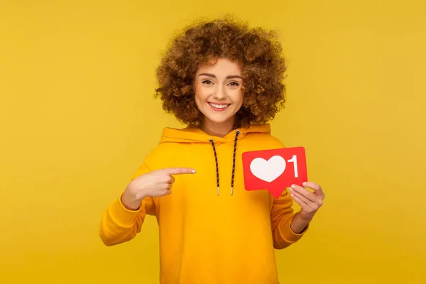stock image Internet blogging. Portrait of joyful curly-haired woman in urban style hoodie pointing at heart like icon, recommending to click on social media button. studio shot isolated on yellow background
