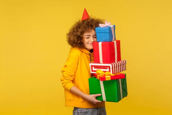 Happy emotions from big mountain of gifts. Portrait of pleased happy curly-haired woman with party cone embracing many present boxes, celebrating birthday. studio shot isolated on yellow background