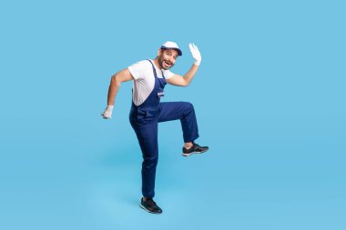 Uzun boylu, mutlu, mavi üniformalı bir tamirci büyük bir adım atıyor, komik bir şekilde gülümsüyor ve aceleyle müşteriye koşuyor. Kurye işi, zamanında teslimat servisi. Stüdyoda çekim izole edildi