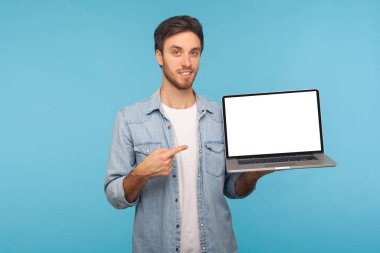 Çalışan kot gömlekli mutlu adamın portresi boş ekranla dizüstü bilgisayarı gösteriyor, ticari görüntü veya metin için boş alan yaratıyor. Stüdyo çekimi mavi arkaplanda izole edildi