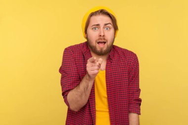 Şapkalı, damalı gömlekli, sakallı, kameraya bakan korkmuş, utangaç ve korkunç bir şey gösteren korkak hippi bir adam. Sarı arka planda izole edilmiş kapalı stüdyo çekimi