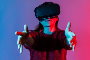 Sanal gerçeklik gözlüklü kız, oyun simülatöründe oynuyor, ellerini uzatmış, sanal dünyada nesneyi tutuyor, VR kulaklık teknolojisi kullanıyor. Renkli neon ışıklı stüdyo çekimi