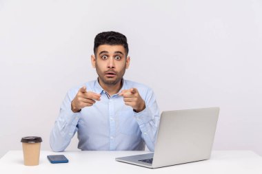 Şaşırmış işadamı, masa başında dizüstü bilgisayarla ofis işyerinde oturuyor, kamerayı işaret ediyor, şaşırmış görünüyor, fark ediyor ve iş arkadaşını işaret ediyor. Beyaz arka planda izole edilmiş kapalı stüdyo çekimi