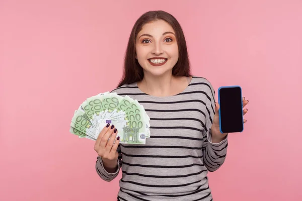 Mobile banking application. Portrait of happy woman in striped sweatshirt holding euro banknotes and cell phone with empty display, mock up for advertise. studio shot isolated on pink background
