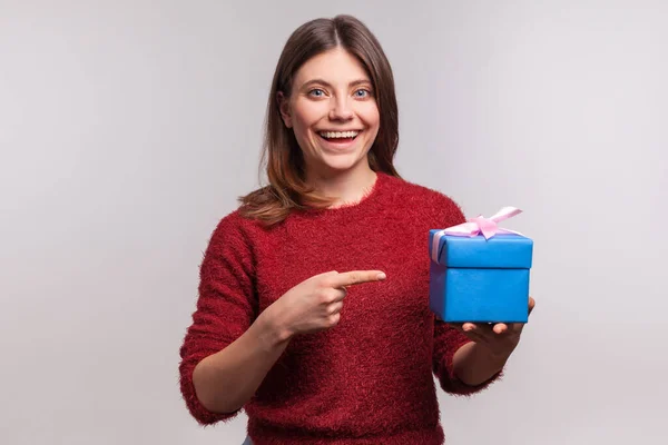 迷人的女孩穿着毛茸茸的毛衣 指着包装精美的礼品盒 面带微笑地看着相机 喜庆生日礼物 庆祝节日 在灰色背景下隔离的工作室拍摄 — 图库照片