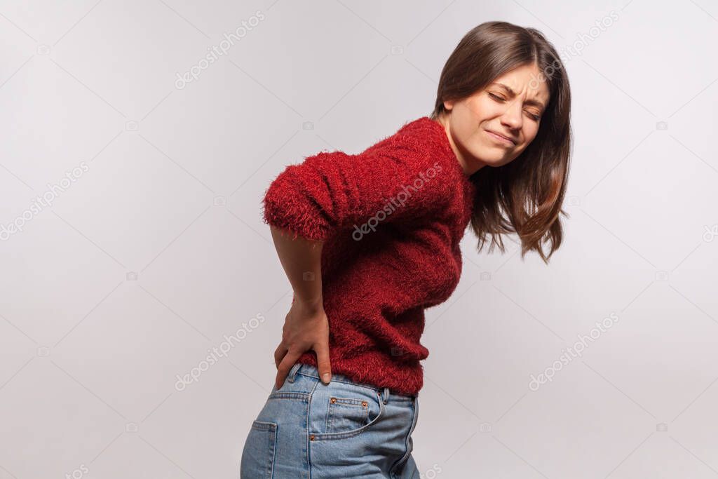 Unhealthy girl in shaggy sweater touching sore back, feeling acute pain, suffering kidney inflammation, pinched nerve, discomfort lower lumbar muscular. indoor studio shot isolated on gray background