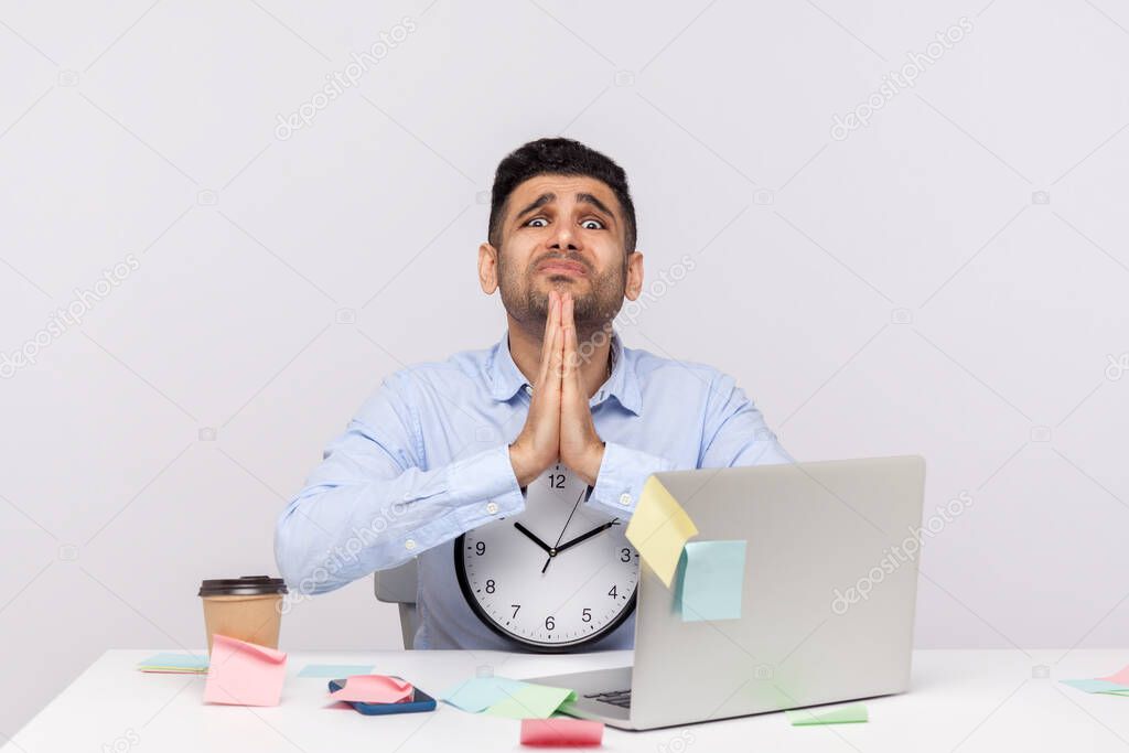 Unhappy man employee sitting in office workplace, holding clock looking imploring with prayer gesture, begging for break, time-out, sticky notes all around reminding of deadline. studio shot isolated