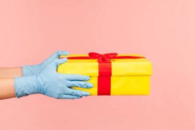 Profil yan görüntüsü: Mavi eldivenli insan eli. Elinde sarı hediye kutusu var. İçerde, stüdyoda, pembe arka planda izole edilmiş..