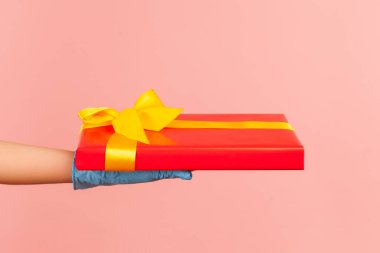 Kırmızı hediye kutusu tutan mavi eldivenli insan elinin profil yan görüntüsü. Paylaşım, verme veya teslim etme kavramı. İçerde, stüdyoda, pembe arka planda izole edilmiş..