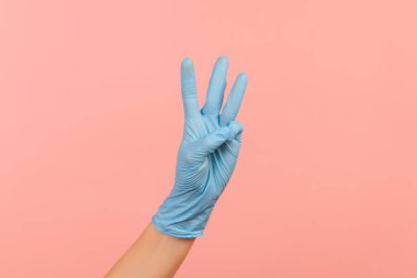 Profil yan görüntüsü. Mavi eldivenli insan eli. 3 numarayı elleri ile gösteriyor. İçerde, stüdyoda, pembe arka planda izole edilmiş..