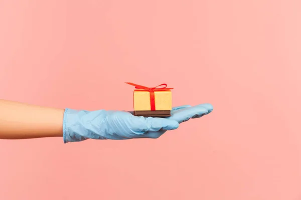 Profile side view closeup of human hand in blue surgical gloves holding small gift box. sharing, giving wedding proposal, or delivery concept. indoor, studio shot, isolated on pink background.