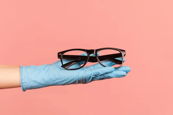 Profile side view closeup of human hand in blue surgical gloves holding and giving black eyeglasses frame. indoor, studio shot, isolated on pink background.