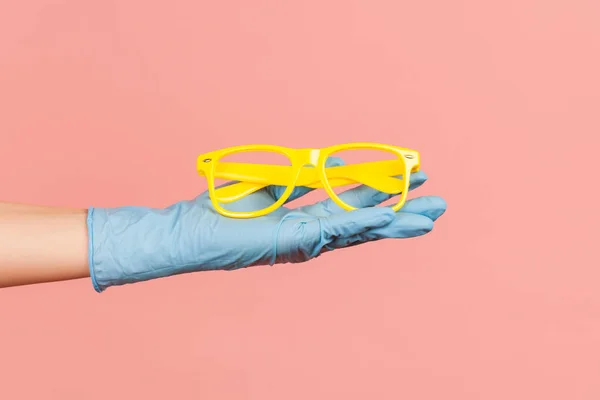 Profile side view closeup of human hand in blue surgical gloves holding and giving yellow eyeglasses frame. indoor, studio shot, isolated on pink background.