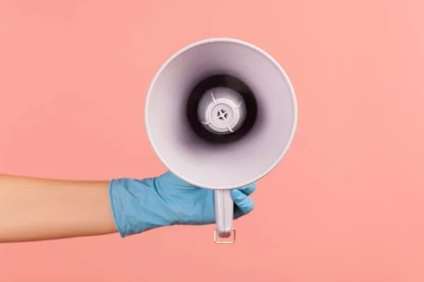 Profile side view closeup of human hand in blue surgical gloves holding megaphone. indoor, studio shot, isolated on pink background.