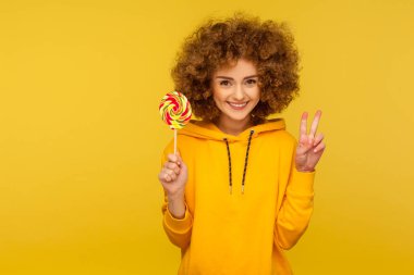 Tatlı gökkuşağı şekeri. Şehir tarzı kapüşonlu mutlu, kıvırcık saçlı kadın portresi lolipop tutuyor ve zafer jesti yapıyor, kameraya gülümsüyor. Stüdyo çekimi sarı arkaplanda izole edildi