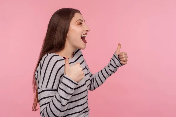 Excellent job, I like it! Side view of joyful excited woman in striped sweatshirt doing thumbs up and shouting in happiness, satisfied with awesome result, successful project. studio shot isolated