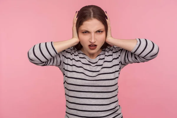 Don\'t want to hear you! Portrait of irritated woman in striped sweatshirt closing ears and looking angry, annoyed by loud sound, unpleasant noise. indoor studio shot isolated on pink background
