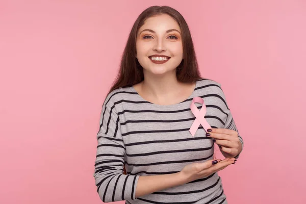 Timely diagnosis, women\'s health care, Portrait of smiling optimistic girl in striped sweatshirt holding pink ribbon, symbol of breast cancer awareness. studio shot isolated on pink background