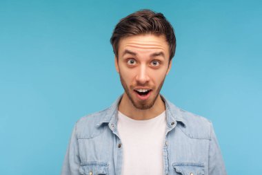 Çalışan kot gömlekli, ağzı açık kameraya bakan, büyük gözlü, beklenmeyen başarıya şaşırmış ve mutlu bir adamın portresi. Mavi arka planda kapalı stüdyo çekimleri