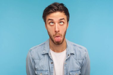 Kot gömlekli aptal bir adamın portresi. Gözleri şaşı, dili dışarı çıkmış, komik, beyinsiz bir yüz ifadesi gösteren komik komik bir surat yapıyor. Mavi arka planda kapalı stüdyo çekimleri