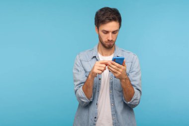 Çalışan kot gömlekli ciddi bir adamın portresi cep telefonuyla sosyal medyada mesaj yolluyor, cep telefonu servislerini kullanıyor, internette sohbet ediyor. Mavi arkaplanda kapalı stüdyo çekimleri, kopyalama alanı