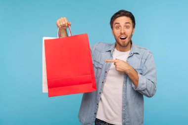 Kırmızı alışveriş torbalarını gösteren kot gömlekli, heyecanlı esmer adamın portresi. Moda mağazasından alınmış paketleri gösteriyor. Stüdyo çekimi mavi arkaplanda izole edildi